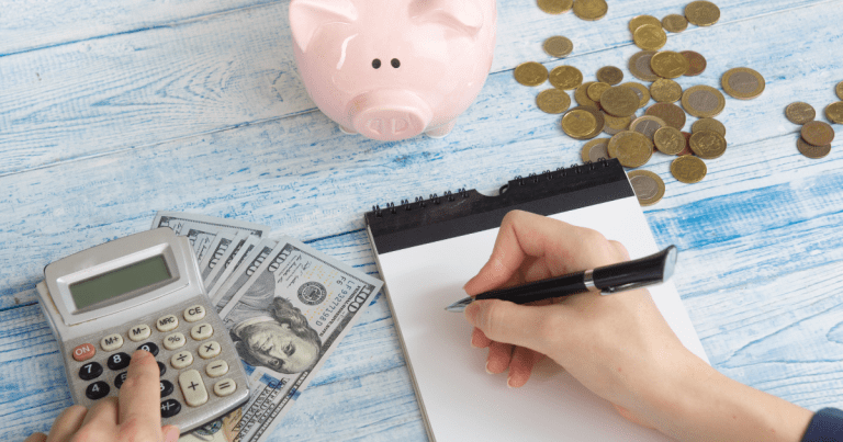 A piggy bank and coins scattered on a table, with a calculator resting on top of bills and a hand holding a pen poised over a notebook. Our accounting firm in Sydney can help you with your personal or business finances.