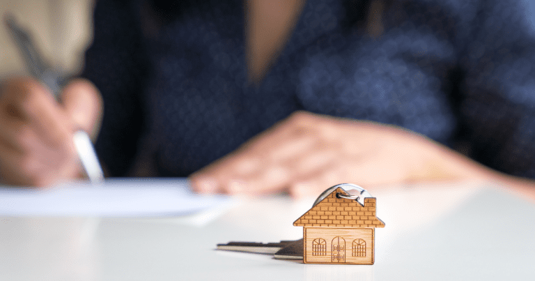 A woman in the background is signing documents, with a miniature house and key on a table in the foreground. Let our small business accountants help you with your financial planning and investment strategies.