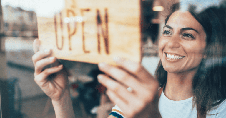 A woman with a wide smile as she attaches an "OPEN" sign. Contact our small business accountants in Sydney for help with business planning and financial management.