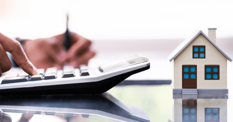 A person writing and using a calculator simultaneously with a miniature house on the table. Our small business accountants can help with your financial planning and management.