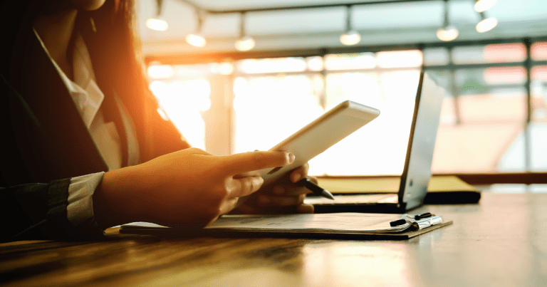 A woman sitting at a table holding a tablet and working on documents, with a laptop also visible on the table. Contact our accounting firm in Sydney for expert bookkeeping and accounting services.