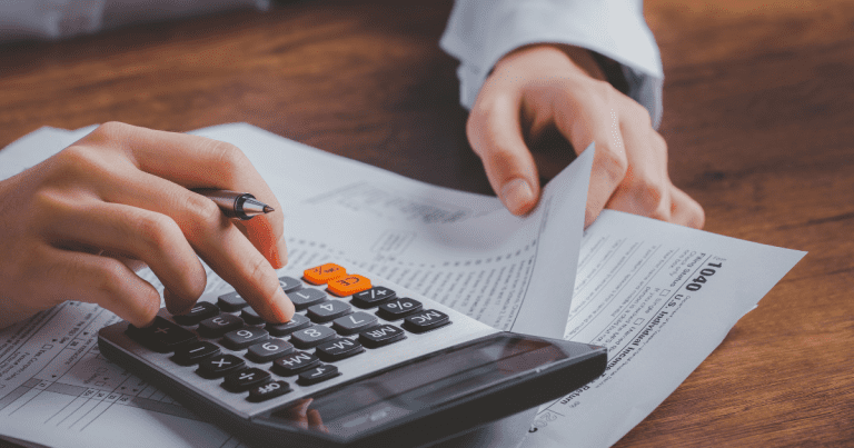 A man is clicking on a calculator while holding some US ITR documents with a pen and notebook beside him, possibly preparing for tax season or financial planning with the help of a tax accountant.