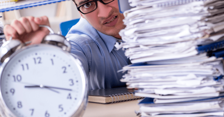 Anxious man holding a clock while facing a pile of documents on his desk, possibly in need of accounting services such as tax or bookkeeping help.