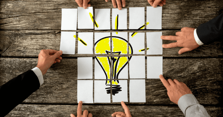 Business accountants' hands gather around and piece together a puzzle of a light bulb on the table in the midst of a brainstorming session.
