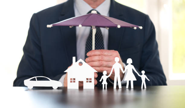 A business accountant holds a miniature umbrella to shelter a paper family, house, and car, symbolising protection and insurance.