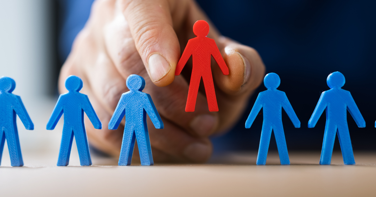 A hand holding a red human figure and about to place it in the centre of a group of blue human figures symbolizing the concept of teamwork and inclusion. Trust our business consultants to guide you in making effective decisions and strategies.