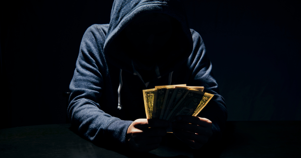 A hooded individual holding dollar bills in a dark setting. Trust our financial experts to help you manage your money wisely.