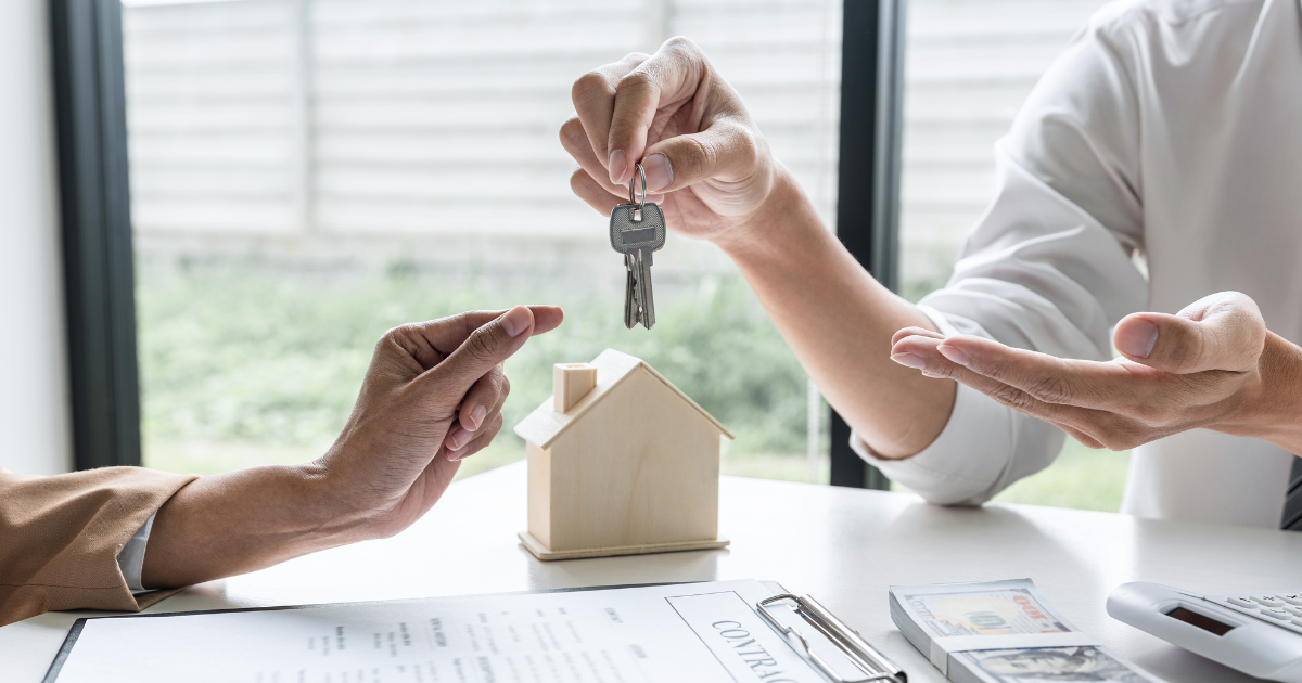 Two individuals are exchanging keys and a contract with a miniature house and dollar bills present on the table, exemplifying the importance of having a trusted business accountant to guide financial decisions and ensure successful transactions.