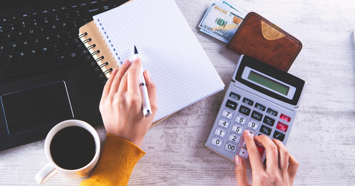 A person holding a pen and calculator, with a notebook, a laptop, a coffee cup, and a wallet with bills visible on top of the table. Our small business accountants can help you stay on top of your finances.