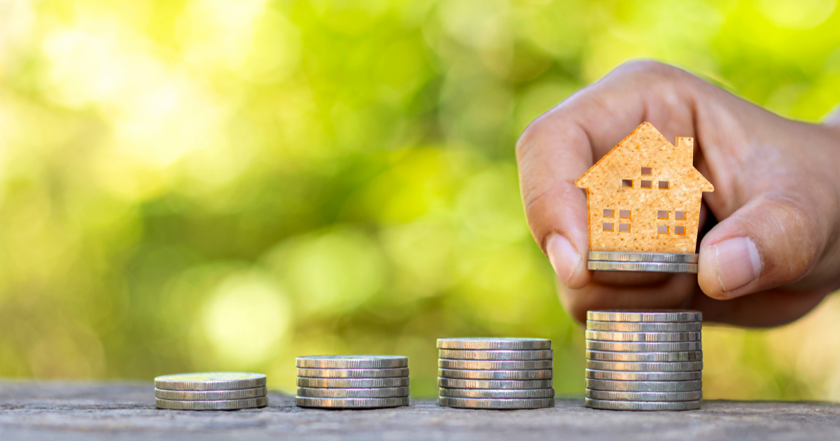 A hand holding a miniature house about to place it on a pile of coins, representing the concept of investing in real estate. Contact our accounting firm in Sydney for expert advice on real estate investing and other investment strategies.